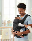 Stork And Baby 4 way baby carrier in black, worn by a father with a baby facing him. Mother stands beside them.