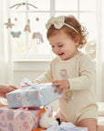 Stork And Baby cream ribbon Alice band with pearls and diamantes worn by a toddler opening gifts in a nursery.