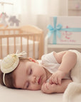 Stork And Baby cream alice band featuring a decorative hat and rose, worn by a sleeping baby in a nursery.