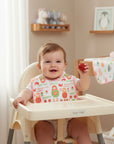 Stork And Baby 100% cotton baby bibs with a doll print and pink trim. A happy baby sits in a high chair holding a strawberry.