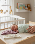 Stork And Baby soft cotton 2-pack girls beanies in mauve and cream with knotted bow detail. A baby sleeps nearby.
