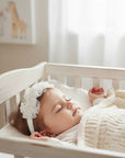 Stork And Baby white lace flower alice band with pearl accents worn by a sleeping baby girl in a nursery crib.