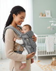 Stork And Baby grey 4-way baby carrier worn by a smiling mother holding her infant in a nursery setting.