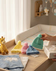 Stork And Baby pastel infant earflap hats in yellow, pink, blue, and mint green with pom-poms.