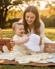 Stork And Baby Mother's Choice white anglaise hat held by a smiling mother for her baby during a sunny park picnic.
