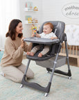 Mother's Choice grey folding baby high chair with a baby sitting in it and a woman kneeling beside it.