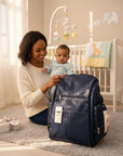 Stork And Baby navy blue faux leather diaper backpack with multiple compartments and side pockets, held by a smiling mother with her baby.