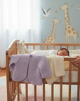 Stork And Baby purple knit cardigan with a single button closure, draped over a crib next to a sleeping infant.