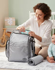 Stork And Baby silver backpack with organized compartments, a changing pad, and a woman packing it next to a baby.