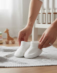 Stork And Baby white 100% cotton infant booties on a grey blanket next to a crib and baby clothes.