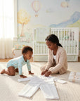 Stork And Baby 5 piece white suit set for infants laid out on a nursery rug with a baby and older girl nearby.
