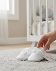 Stork And Baby white embroidered christening booties for baby boys with lace-up detail on a soft nursery rug.