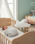 Stork And Baby white cloud plush pillow with "baby" embroidered, held over a crib with a sleeping infant.