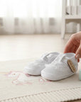 Stork And Baby white christening booties with T-bar strap and delicate lace trim on a unicorn-patterned baby blanket.