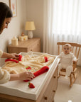 Stork And Baby girls coral fleece tracksuit in cream with red trim and strawberry embroidery. A baby sits in a rocking chair in the background.