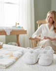 Stork And Baby Mothers Choice white lace christening booties with ribbon ties on a nursery bed with a woman nearby