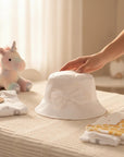 Stork And Baby white satin floppy hat with a bow for baby girls, displayed on a nursery table with baby clothes.