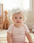 Baby girl wearing a Stork And Baby white satin and lace bow Alice band in a nursery setting.
