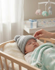 Stork And Baby grey knit pom pom beanie on a sleeping baby in a crib.