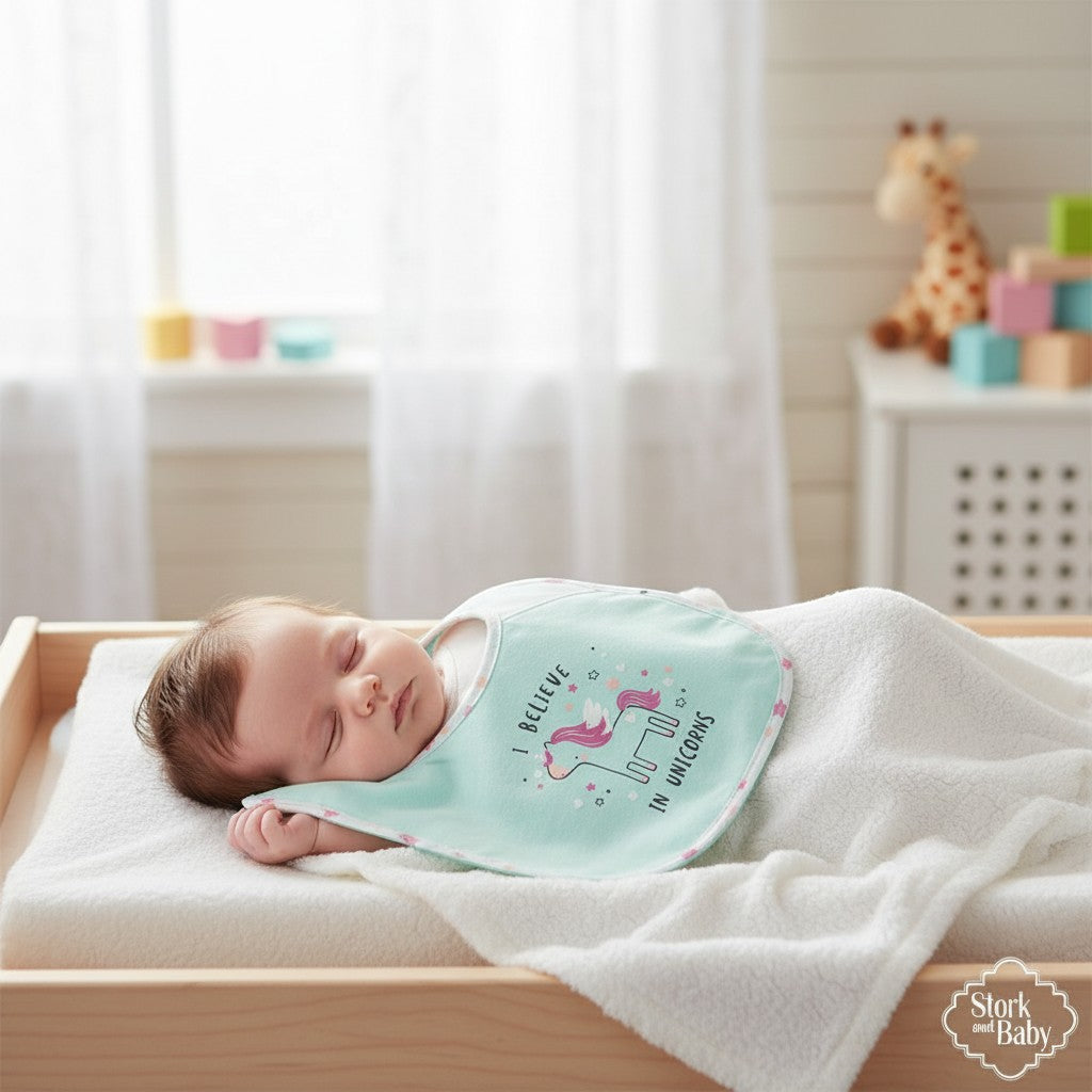 Stork And Baby infant's light blue bib with a pink unicorn graphic and 'I Believe In Unicorns' text.