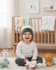 Stork And Baby infants winter hat in grey and white stripes, worn by a smiling baby sitting on the floor.