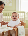 Stork And Baby white embroidered linen/cotton bibs on a smiling baby in a high chair, with a mother feeding.