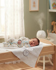 Stork And Baby Mothers Choice white knitted shawl draped over a changing table with a sleeping baby in a nursery.