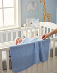 Stork And Baby Mothers Choice blue cable-knit baby shawl in a nursery cot with a sleeping newborn.
