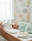 Stork And Baby Mother's Choice white satin baby cap held by a mother next to a sleeping newborn in a nursery