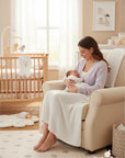 Stork And Baby Mothers Choice white acrylic baby shawl draped over a mother holding her newborn in a nursery chair.