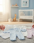 Stork And Baby pastel babygrows in pink, blue, and white. 100% cotton, long-sleeved with snap closures. A sleeping baby lies nearby.