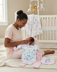 Stork And Baby 'Rainbow' 3-piece baby set with tie-dye bodysuit, pink leggings, and rainbow bib. Mother holding baby.