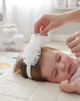 Stork And Baby white lace flower alice band with pearl detail on a sleeping baby wearing a pink unicorn onesie.