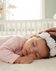 Stork And Baby infant white lace headband featuring three decorative roses, worn by a sleeping baby in a nursery.